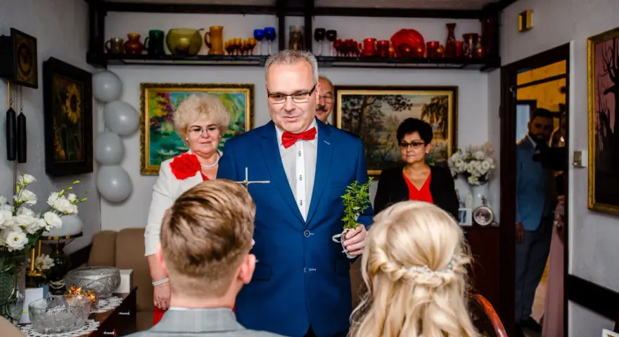 Mężczyzna w niebieskiej marynarce dokonuje tradycyjne błogosławieństwo, trzymając krzyż i gałązkę.